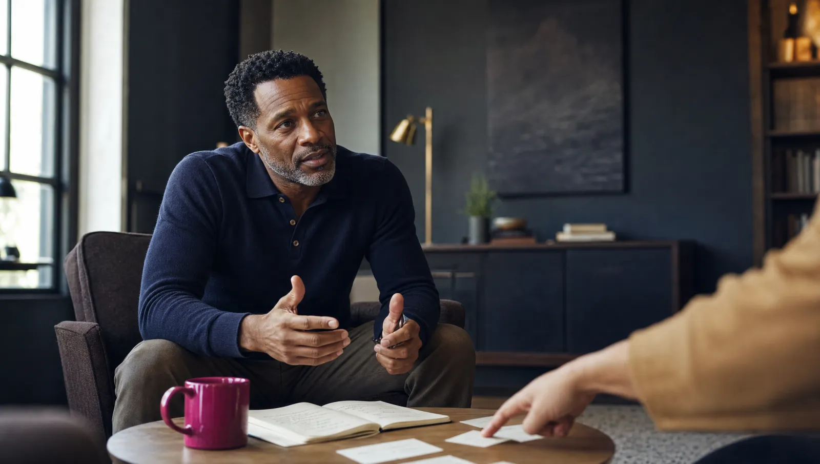 A man in his forties leans forward in conversation, working through handwritten notes and cards spread on the table with someone guiding him.