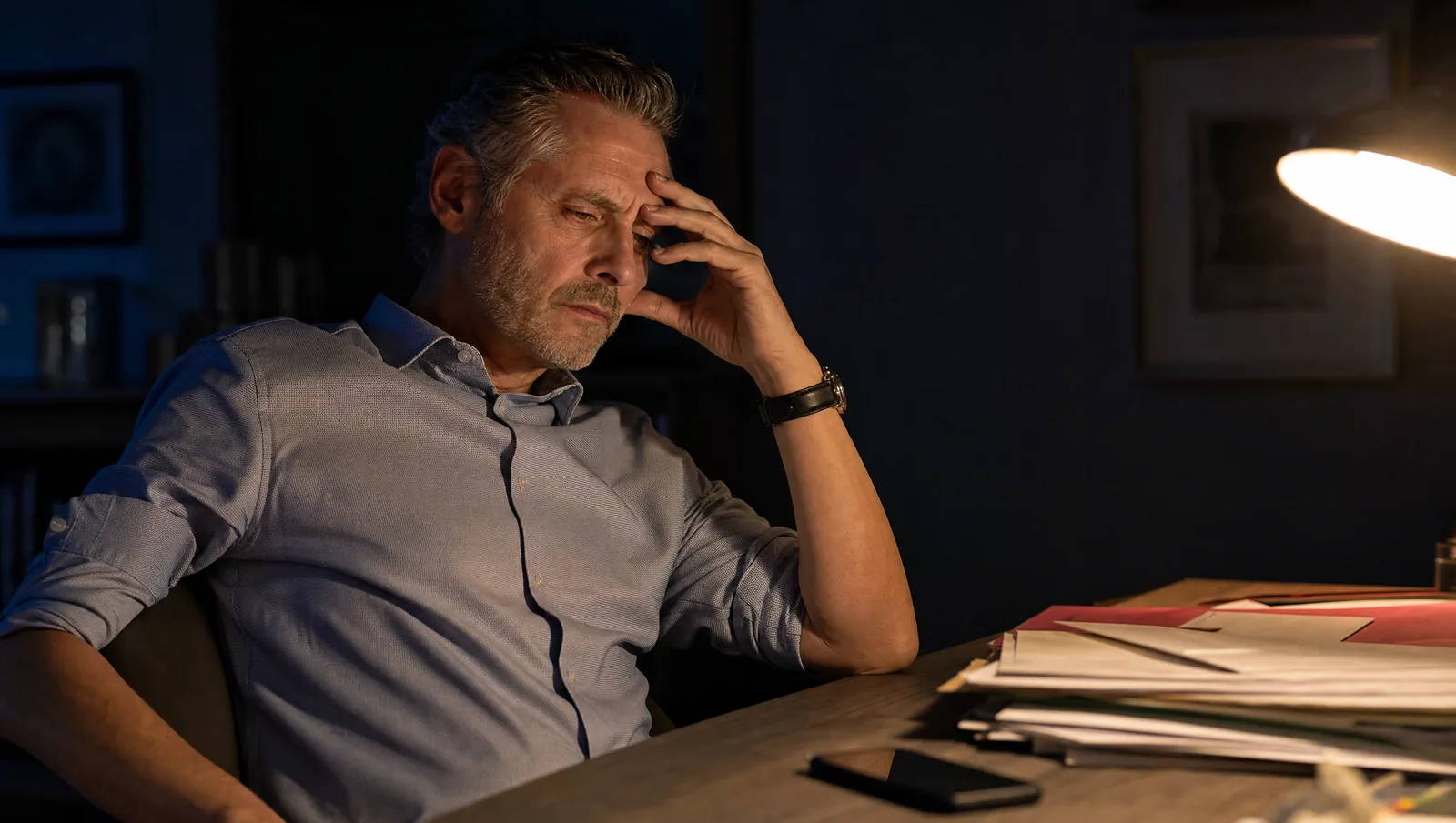 A senior professional at his desk at night, hand to his brow beside a stack of applications and a silent phone