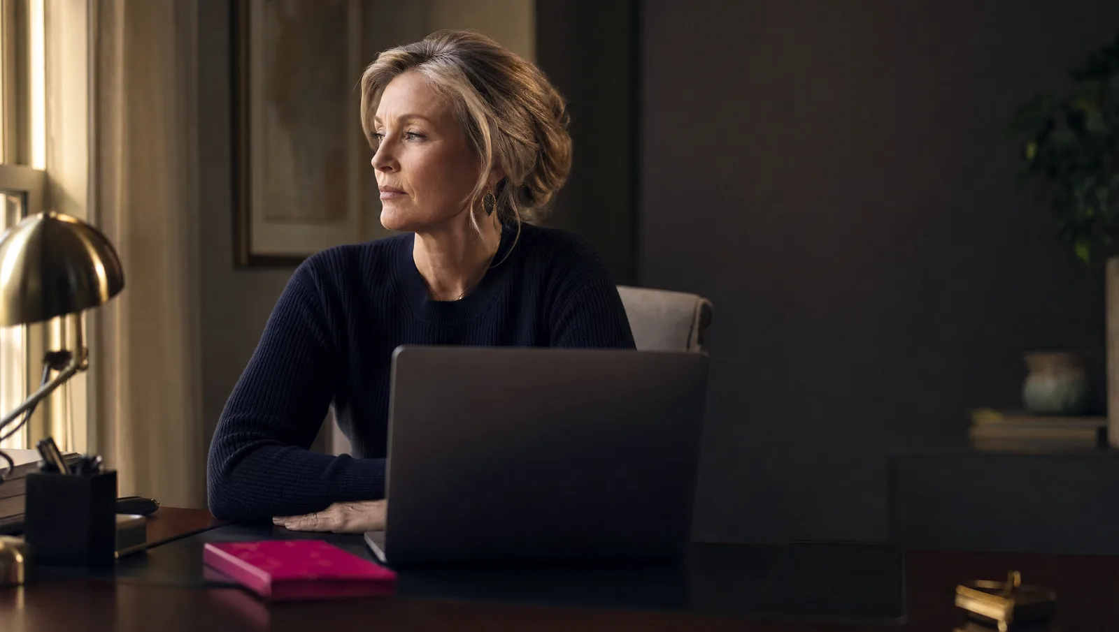 A senior professional pauses at her desk, gazing past her laptop out of the window, quietly unfulfilled