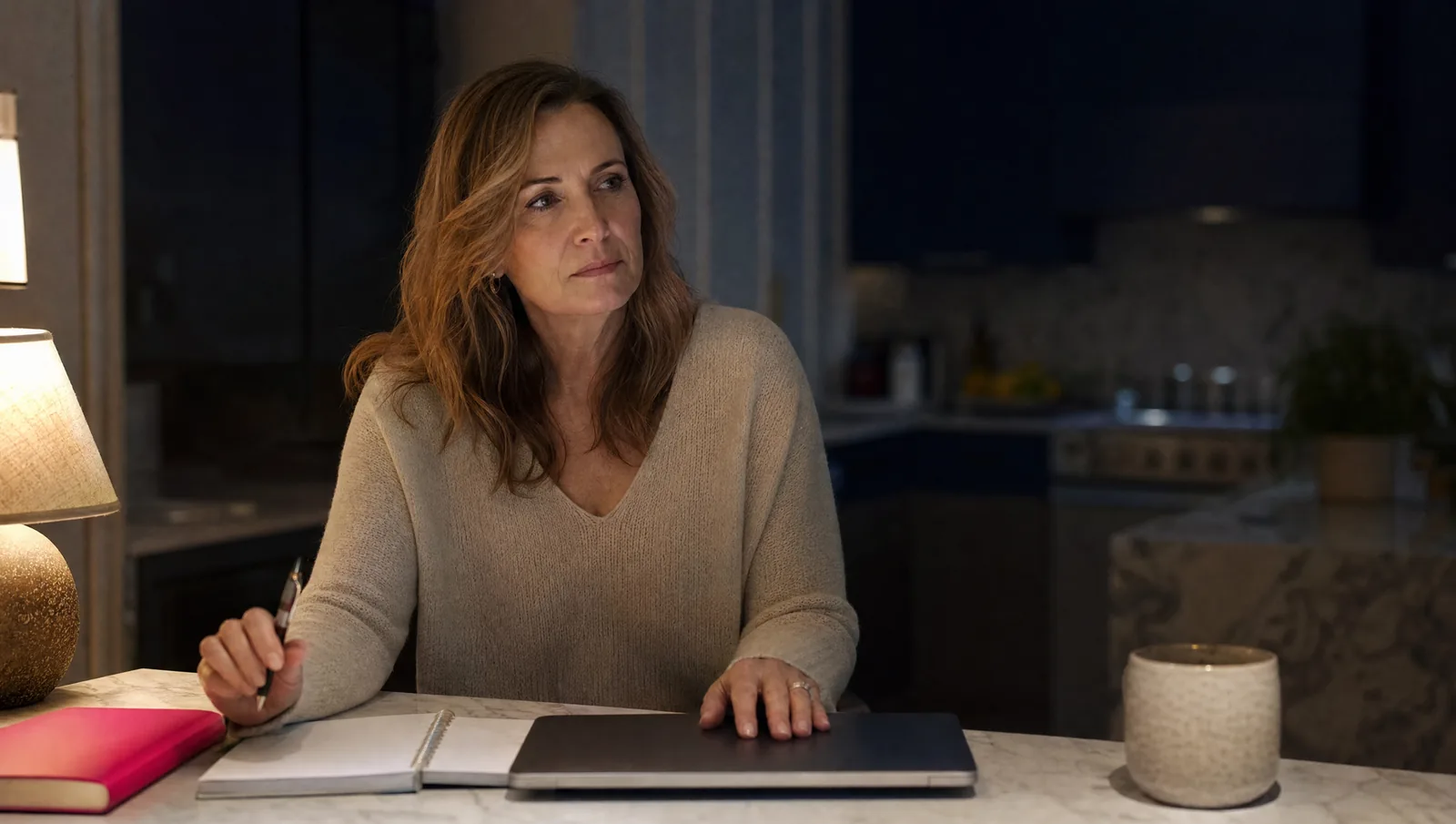 A woman at her kitchen island late at night, hand on her closed laptop, pen over an untouched notebook, deciding